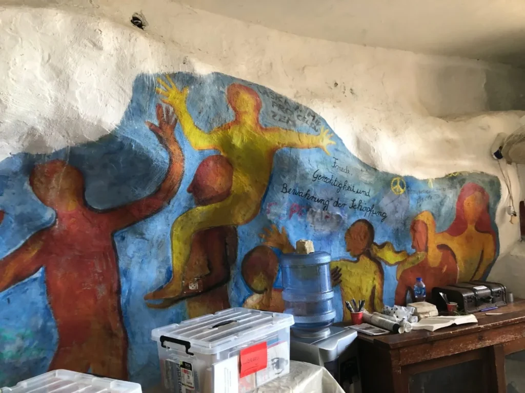 A mural of multicolored human silhouettes is painted on a curved wall above storage boxes, a water cooler, and office supplies in an indoor space.
