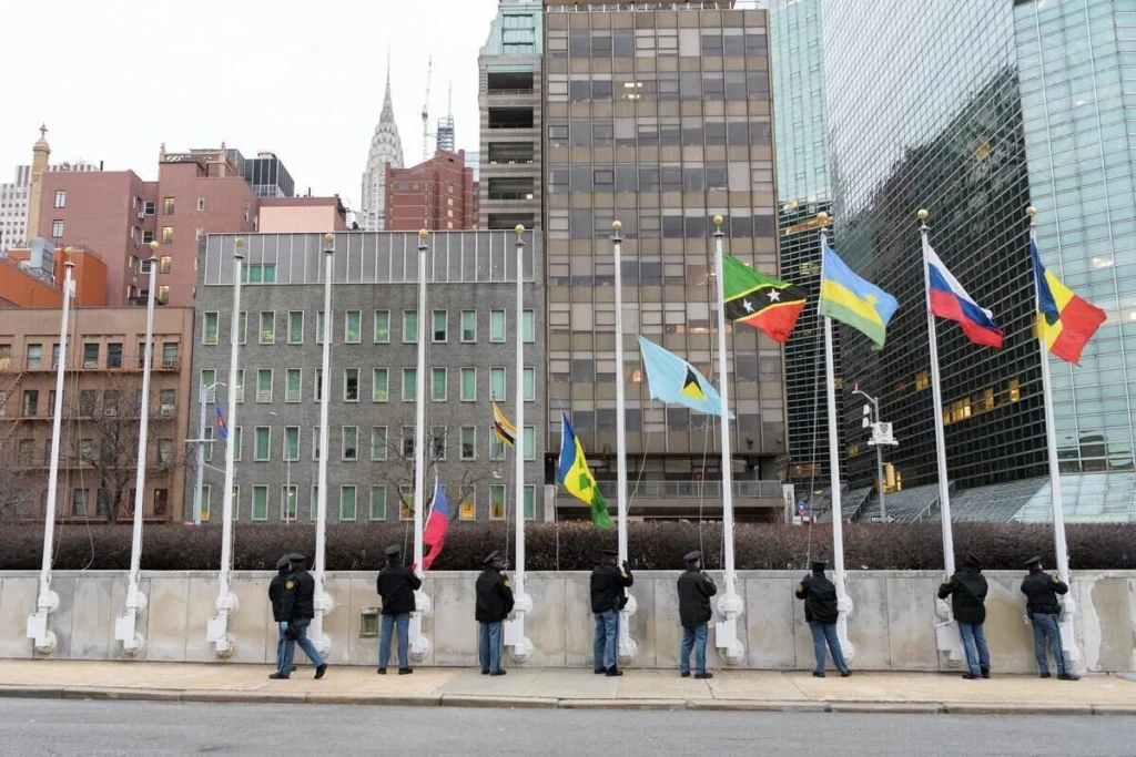 Several people lower national flags outside a modern office building, with city skyscrapers visible in the background.