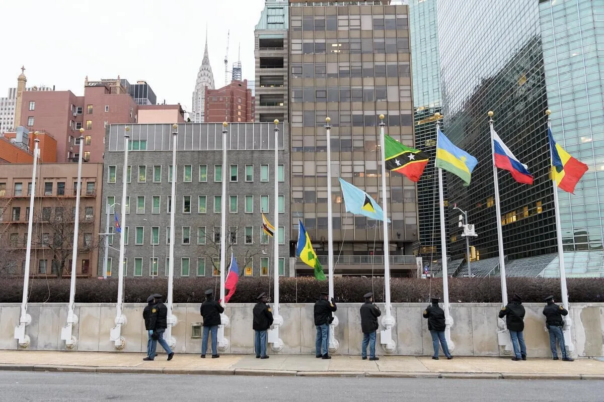 UN Photo/Manuel Elias Several people lower national flags outside a modern office building, with city skyscrapers visible in the background.